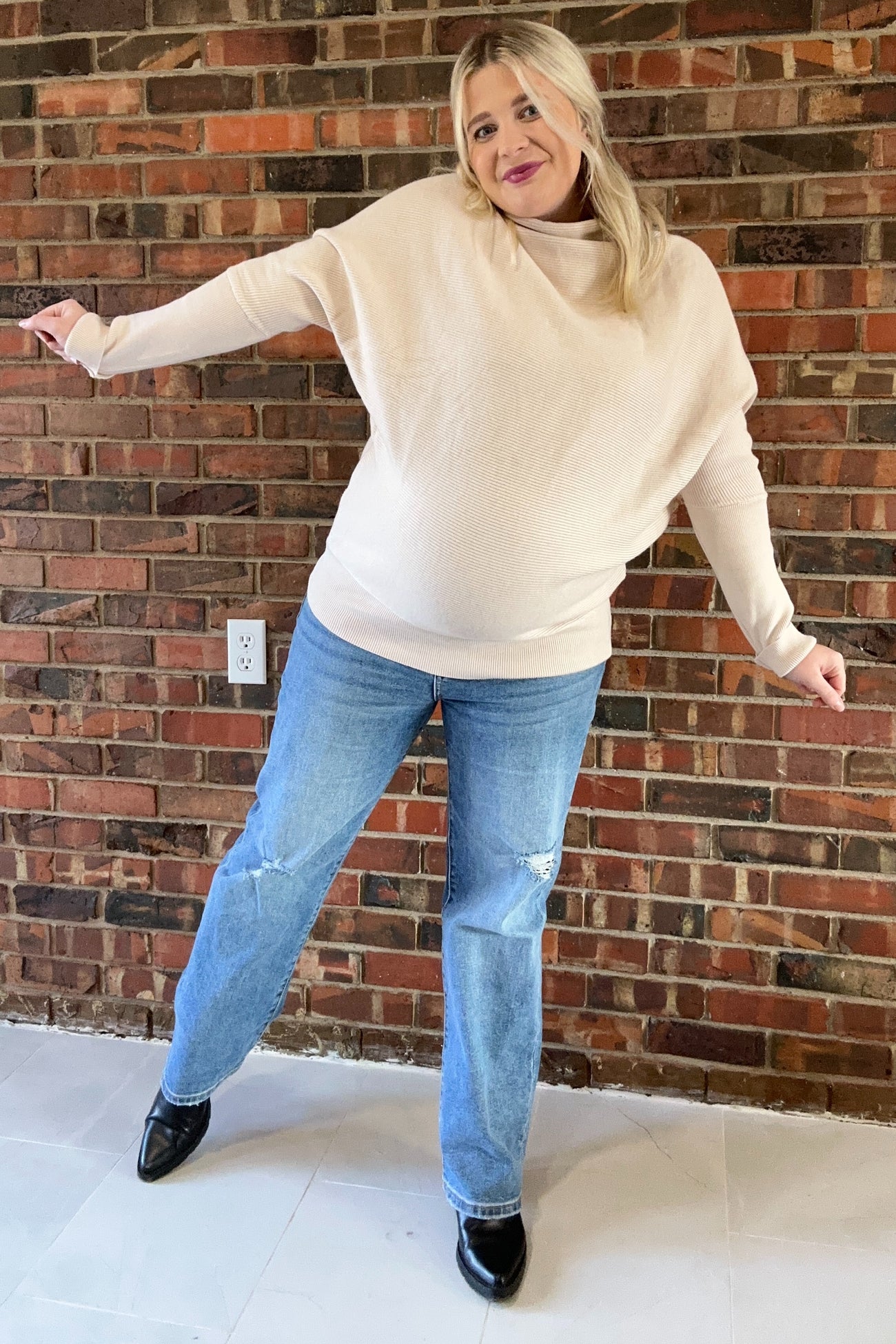 woman wearing a beige oversized maternity sweater and blue distressed jeans, showing her baby bump