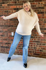 woman wearing a beige oversized maternity sweater and blue distressed jeans, showing her baby bump