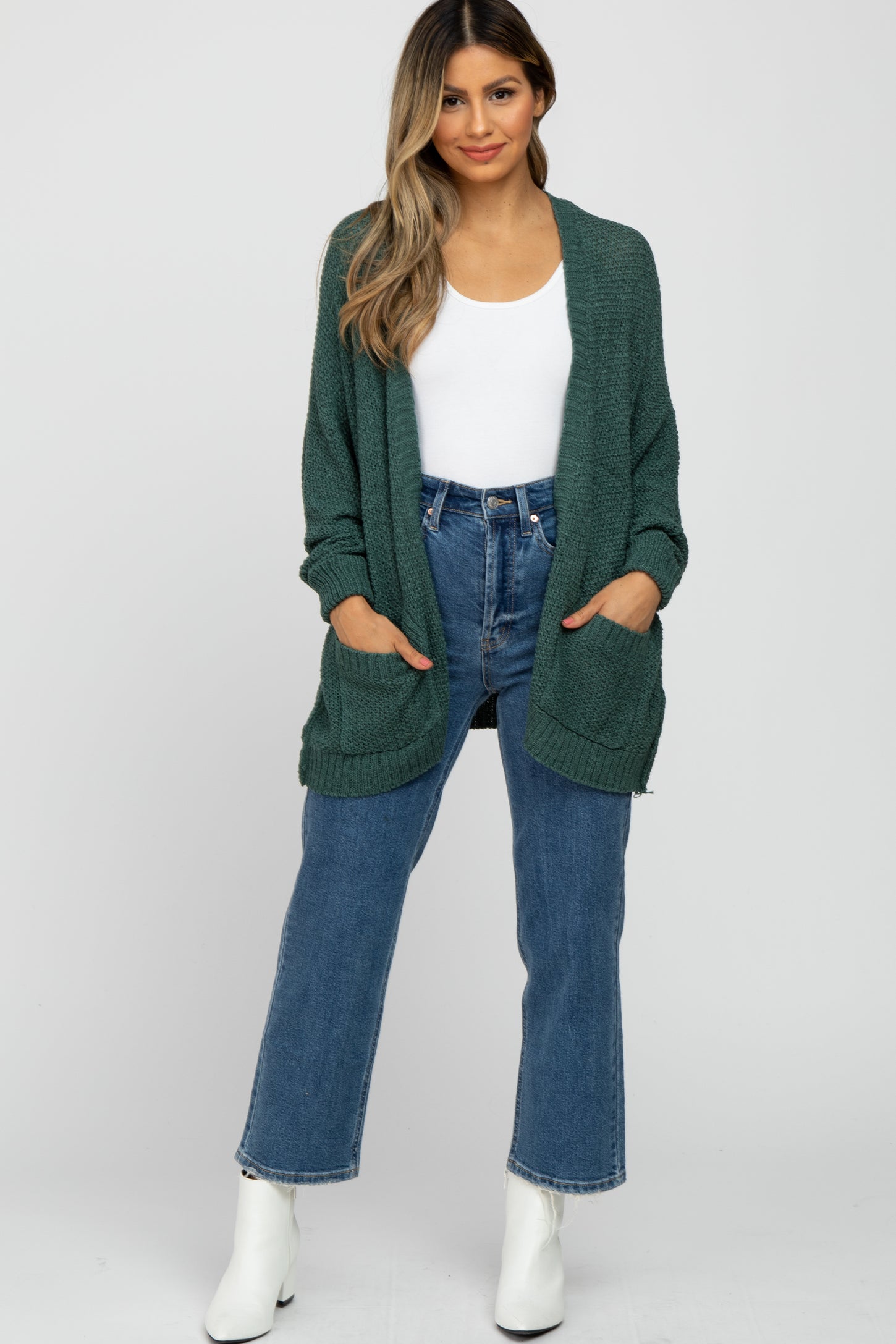 woman wearing a dark green knitted cardigan over a white top and high-waisted blue jeans with white ankle boots