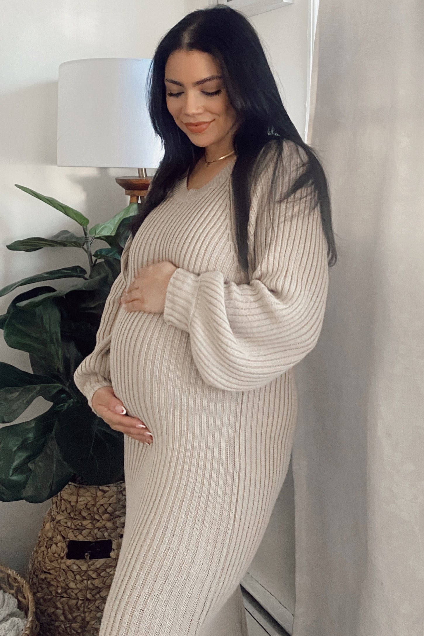 woman wearing a beige knit maternity dress with long sleeves and a fitted silhouette, holding her baby bump