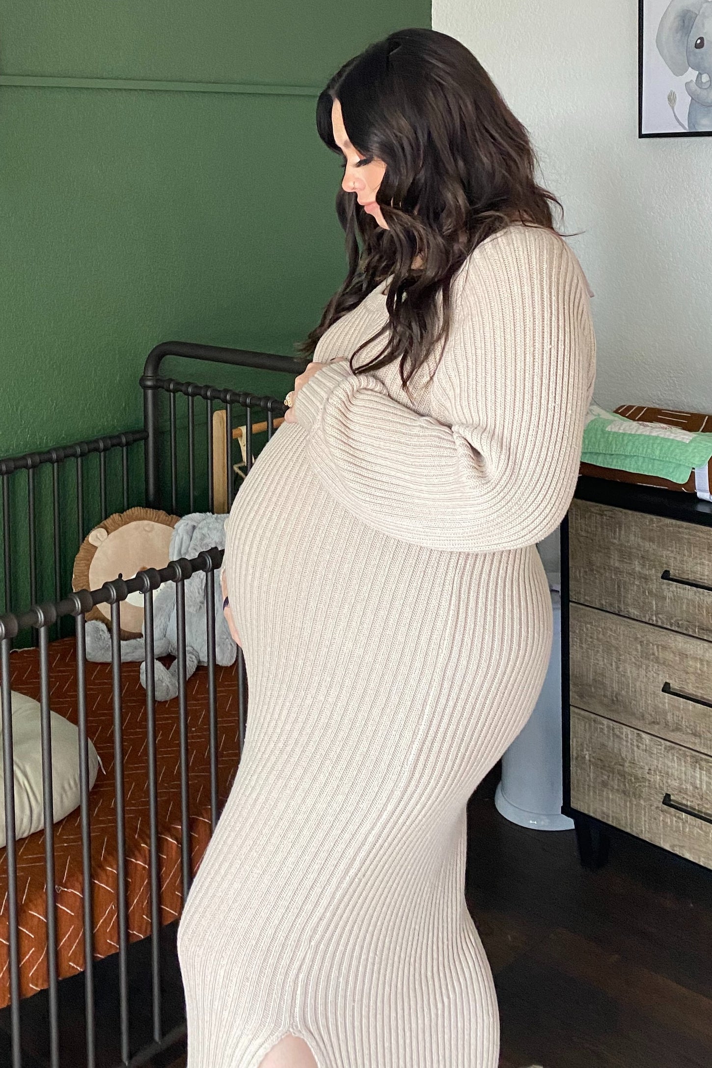 woman wearing a beige ribbed maternity dress highlighting her baby bump while standing beside a crib