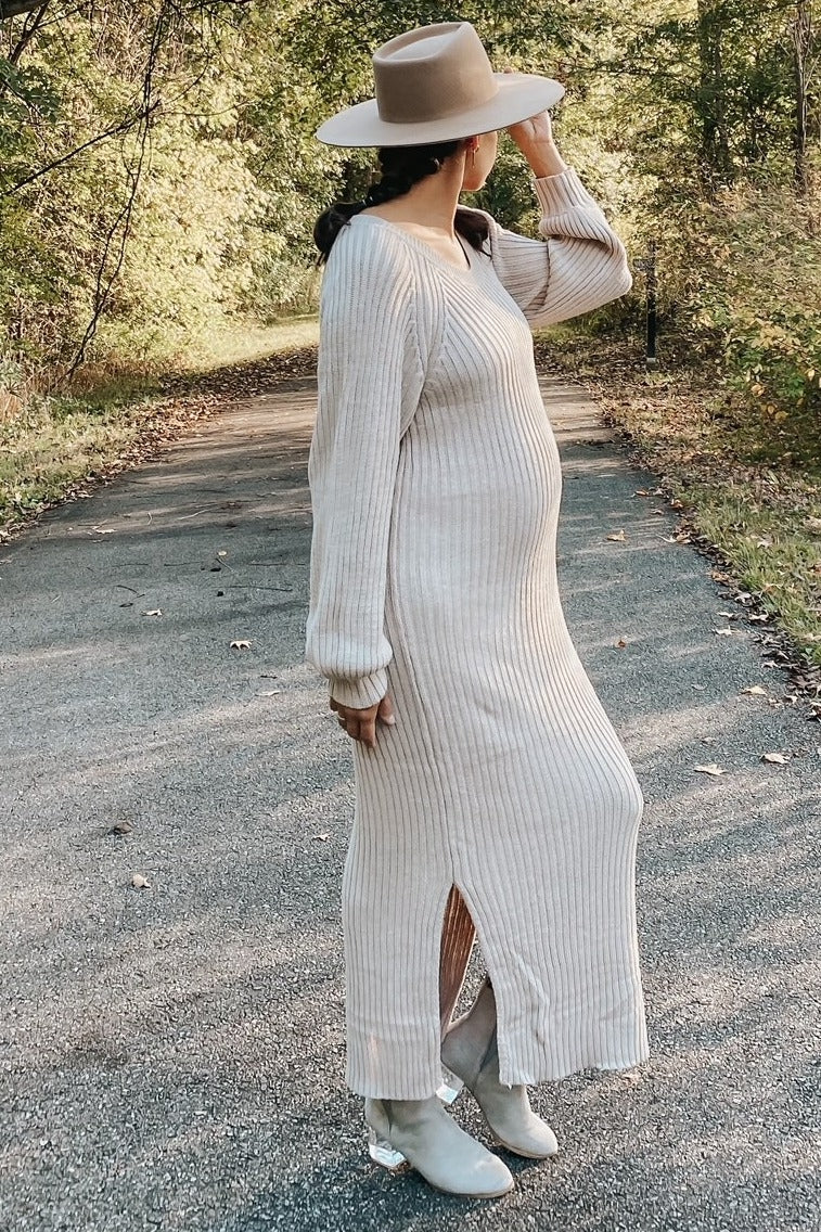woman wearing a beige ribbed maternity dress with side slits and long sleeves, holding her hat, showcasing her baby bump