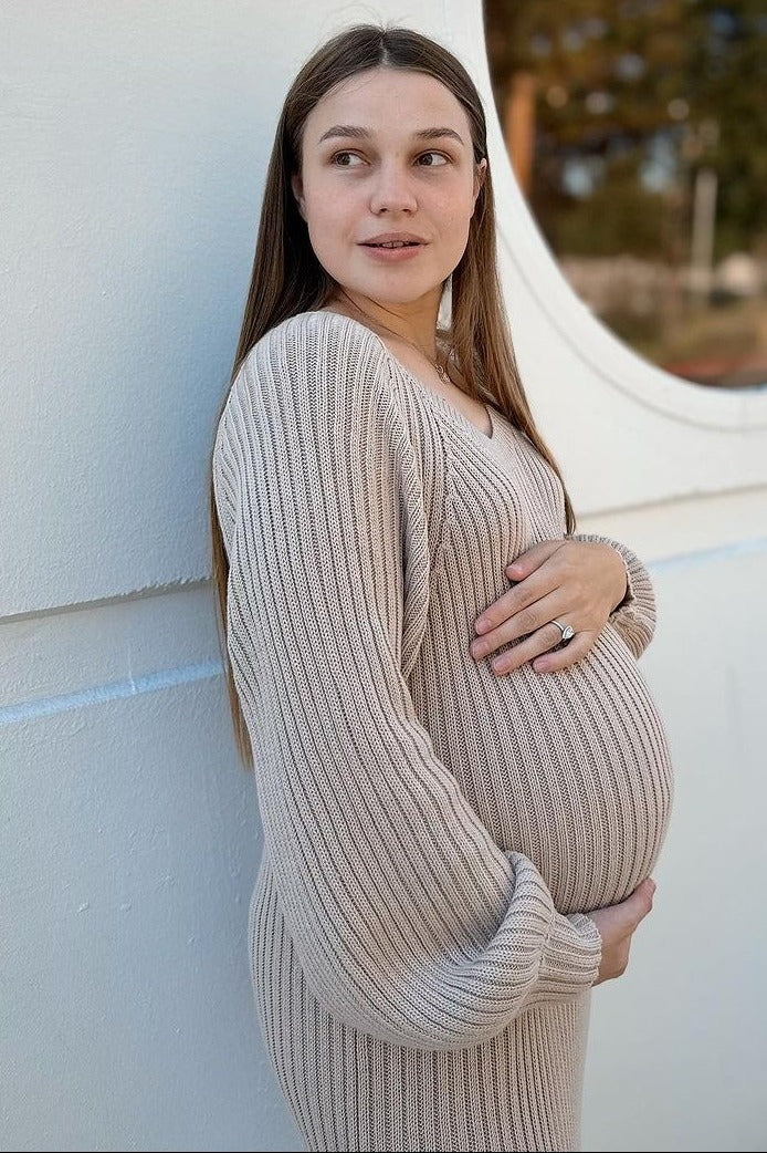 woman wearing a light beige ribbed maternity sweater dress, holding her baby bump and standing against a white background