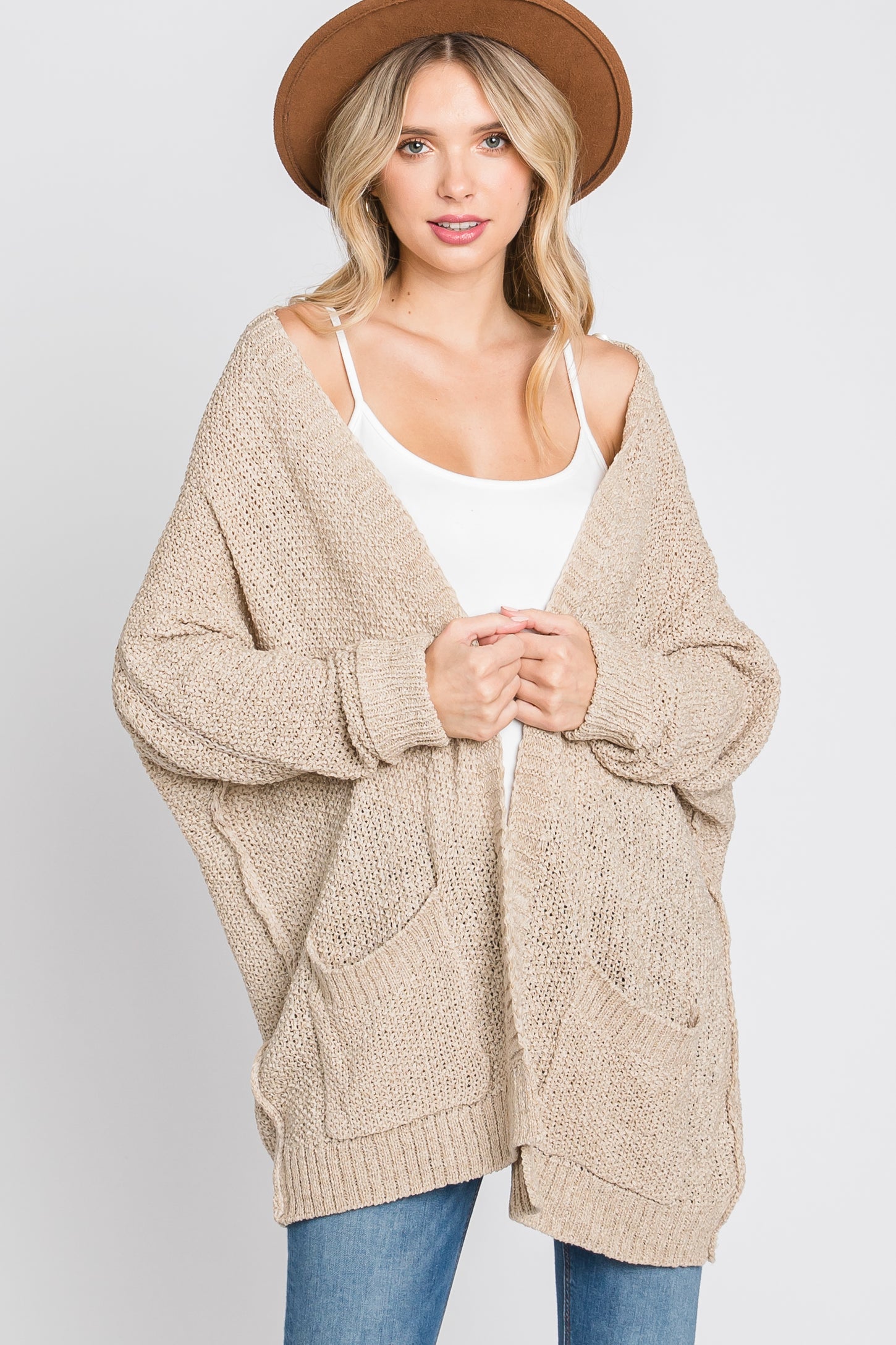 woman wearing a beige oversized knit cardigan with pockets and a white tank top, styled with a brown hat