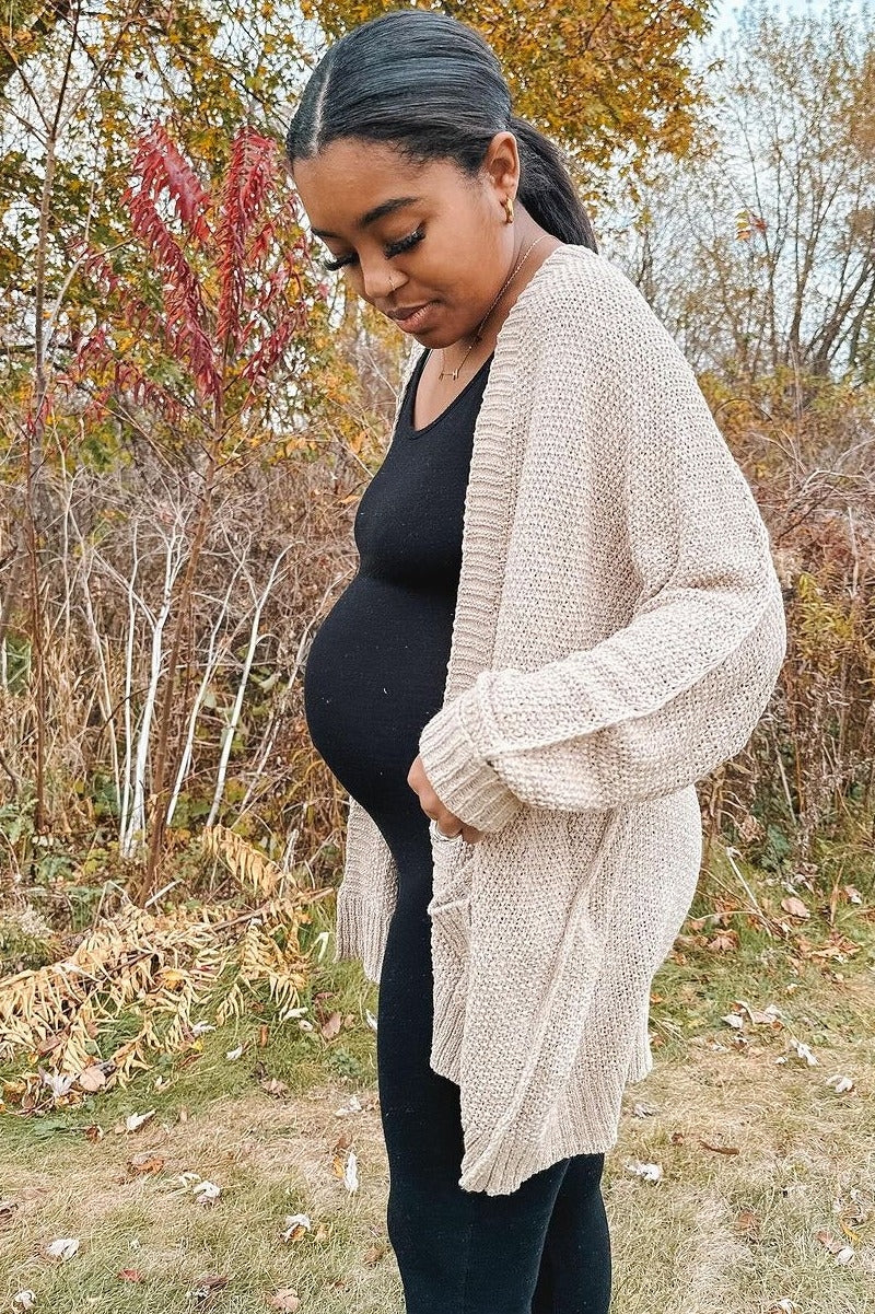 woman wearing a beige knitted maternity cardigan over a black dress, showing a visible baby bump