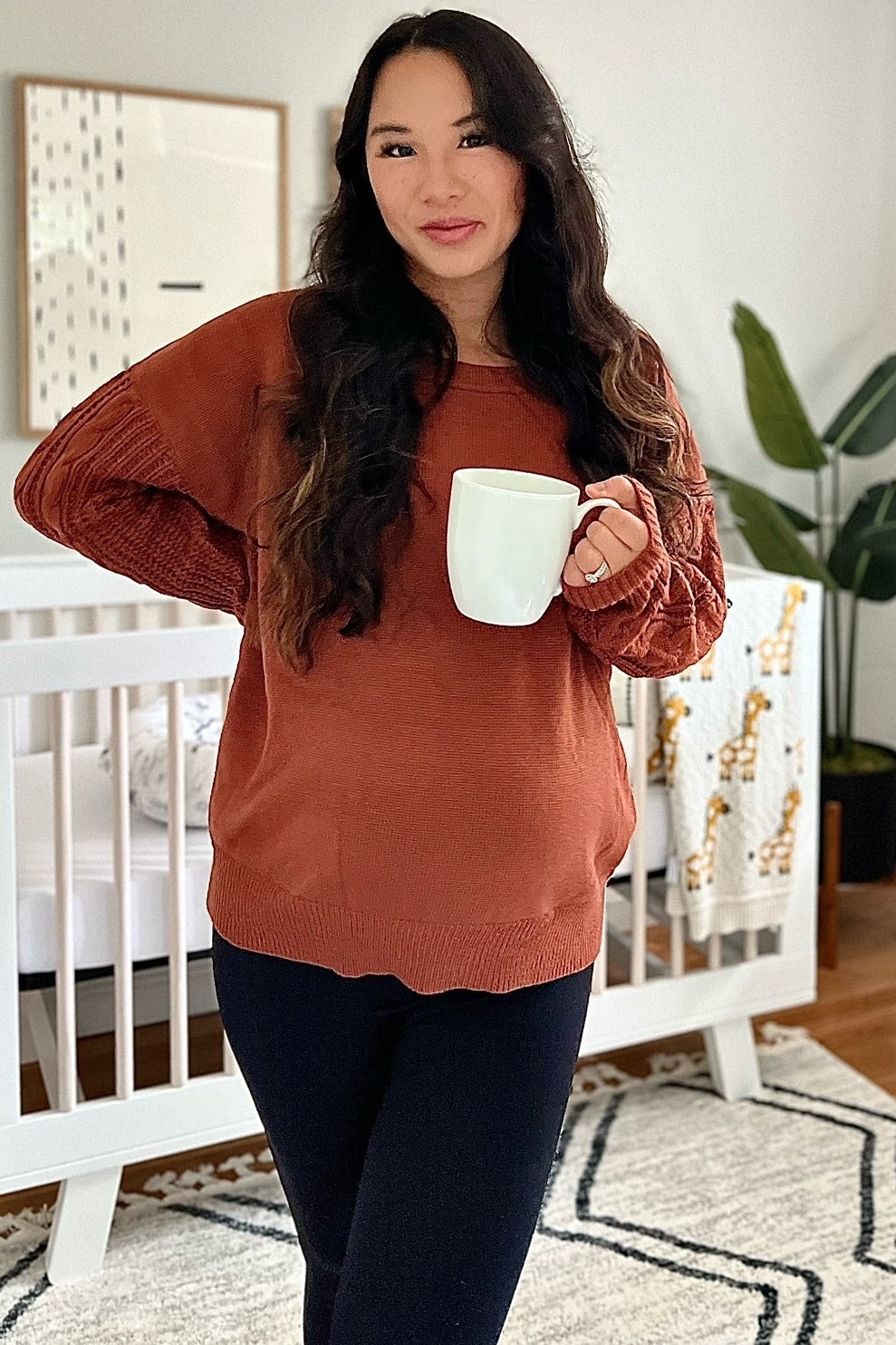 woman wearing a rust-colored maternity sweater with textured sleeves, holding a white mug and showing her baby bump