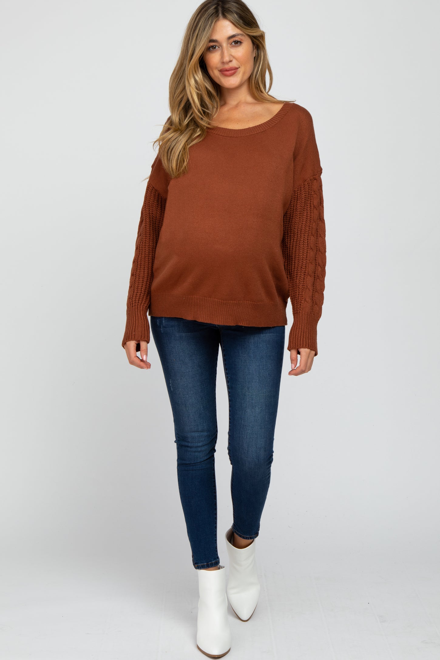 woman wearing a rust cable knit sleeve maternity sweater and blue skinny jeans, with a visible baby bump