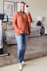 woman wearing an orange cable knit maternity sweater and holding her baby bump, paired with blue distressed jeans