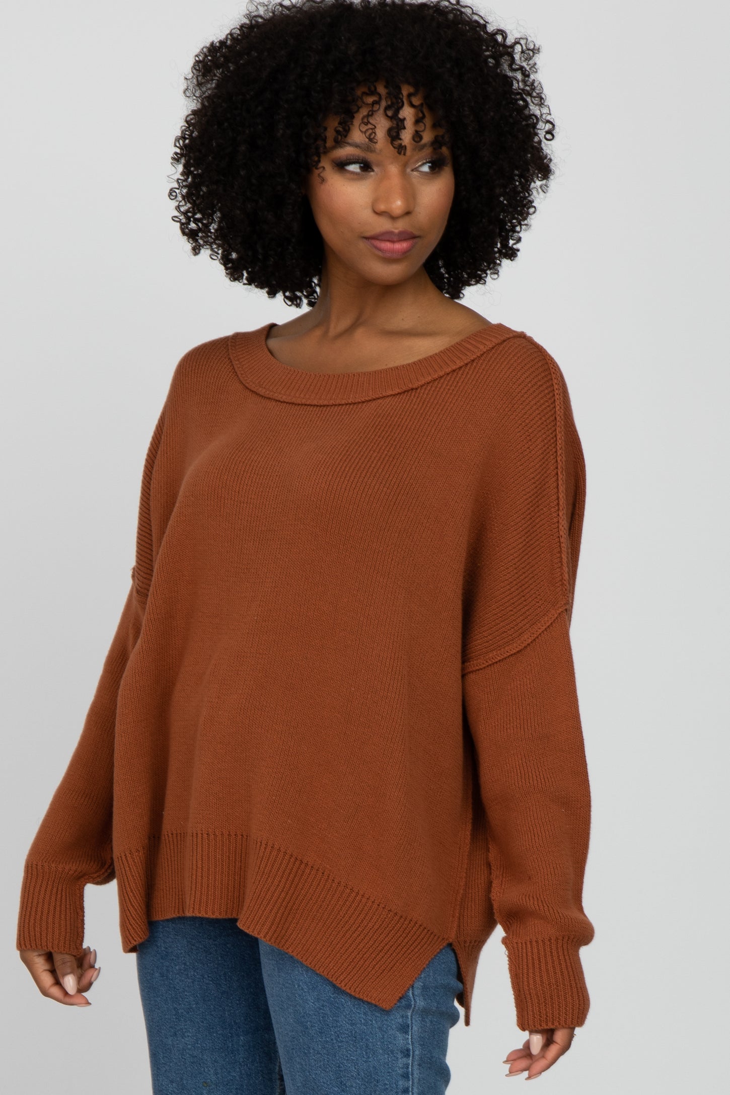 woman wearing a rust oversized sweater with a wide neckline and side slits, paired with blue jeans