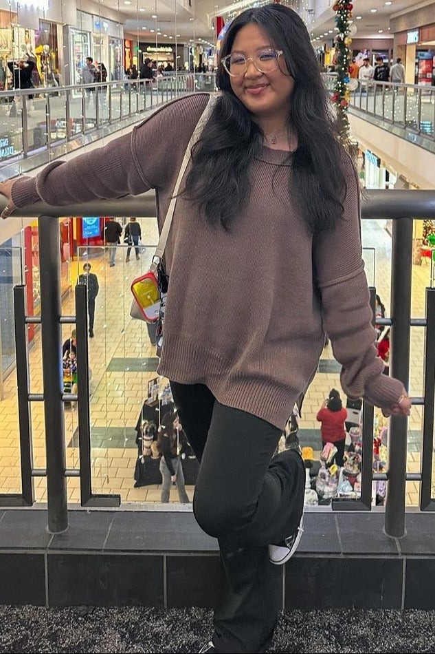 woman wearing a brown oversized sweater and black flared pants, posing with one hand on a railing in a shopping mall