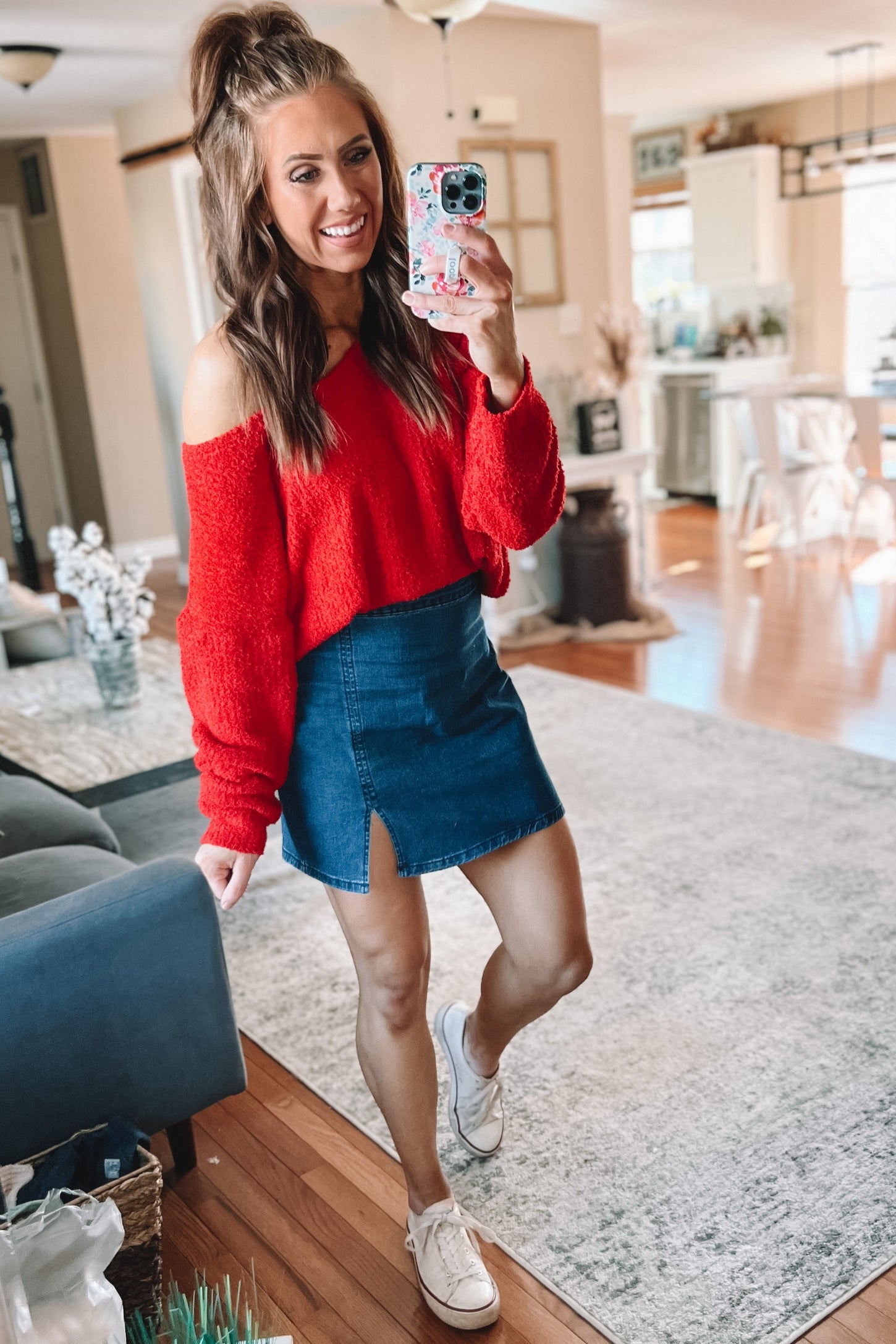 woman wearing a red off-the-shoulder sweater and a blue denim mini skirt, standing in a home interior