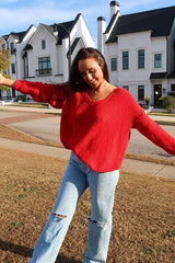 woman wearing a red oversized sweater and light blue distressed jeans, smiling outdoors with arms outstretched