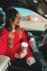 woman wearing a red sweater and patterned leggings, sitting in a car and holding a coffee cup