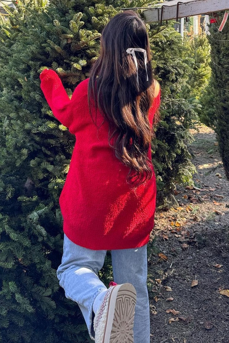 woman wearing a slouchy red sweater and light blue jeans standing next to a tree