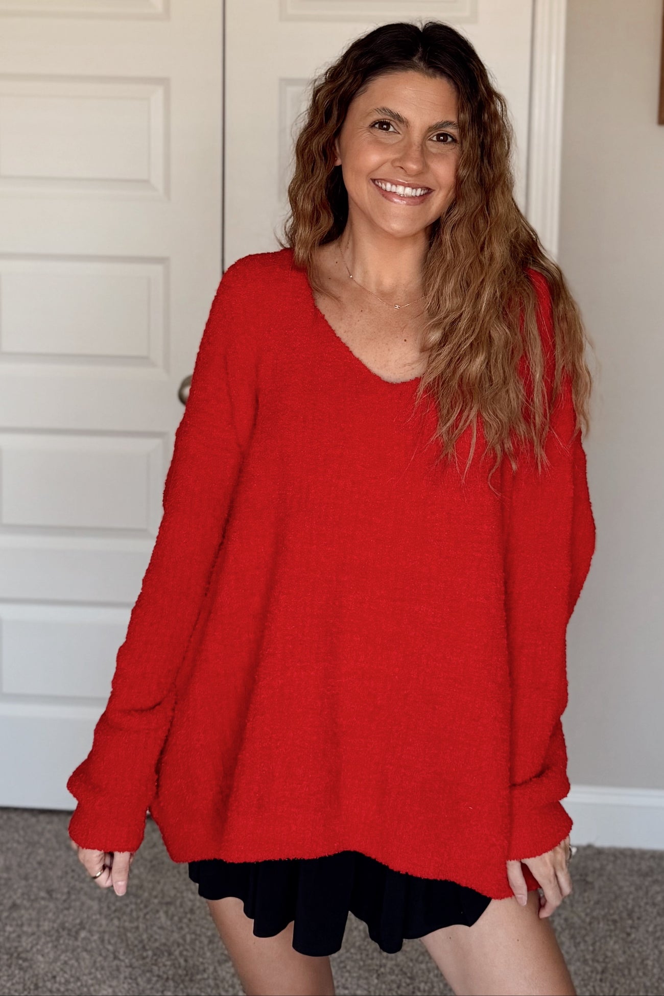 woman wearing a bright red oversized sweater with a v-neckline and black shorts, smiling at the camera
