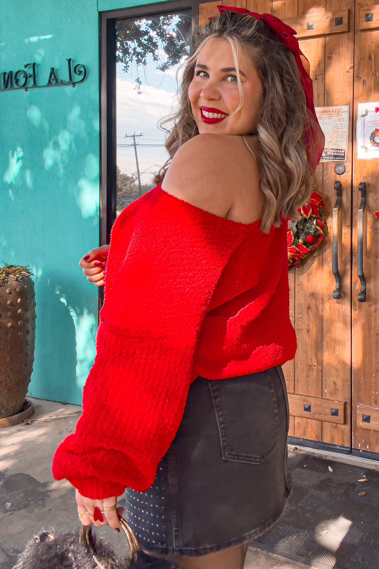 woman wearing a red off-the-shoulder sweater and a black denim skirt, smiling with a red bow in her hair
