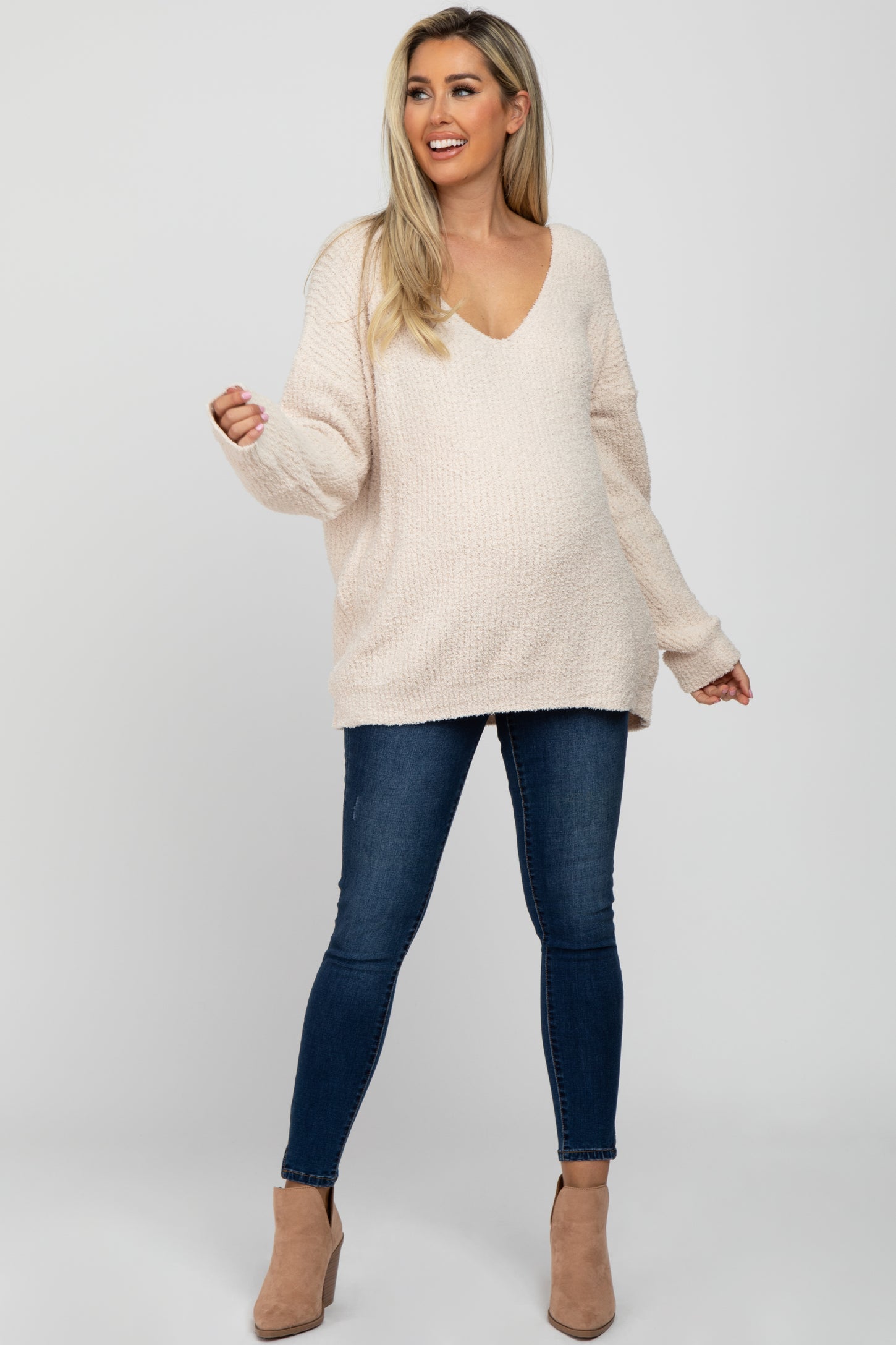 woman wearing a light beige oversized maternity sweater with a v-neck and distressed blue maternity jeans, showing her baby bump