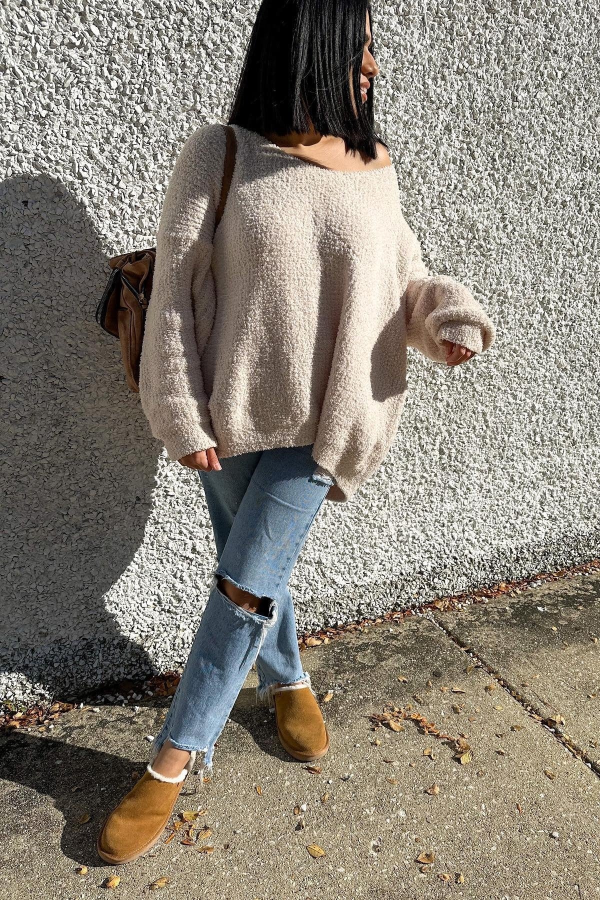 woman wearing a cozy beige oversized sweater, distressed jeans, and tan slip-on shoes, standing against a textured wall