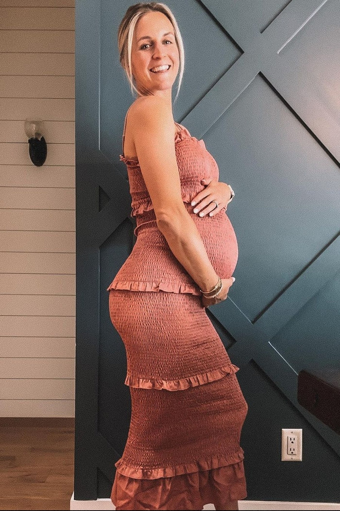 woman wearing a rose-colored maternity dress with ruffles, showing her baby bump and standing against a patterned wall