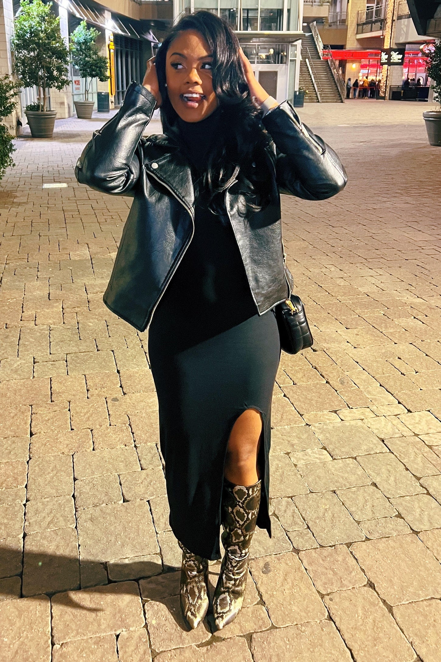 woman wearing a black dress with a thigh-high slit, black leather jacket, and snake print knee-high boots