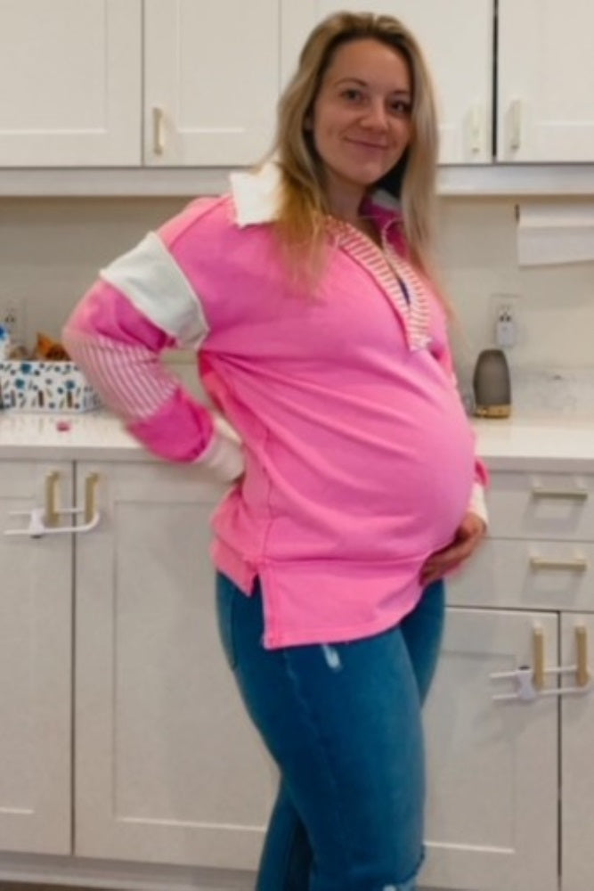 woman wearing a bright pink maternity pullover with a collar, holding her baby bump, paired with blue skinny jeans