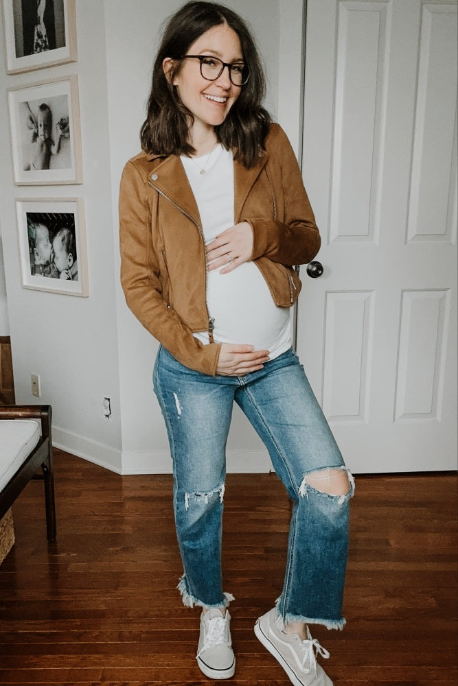 woman wearing a brown suede maternity jacket, white t-shirt, distressed jeans, and holding her baby bump