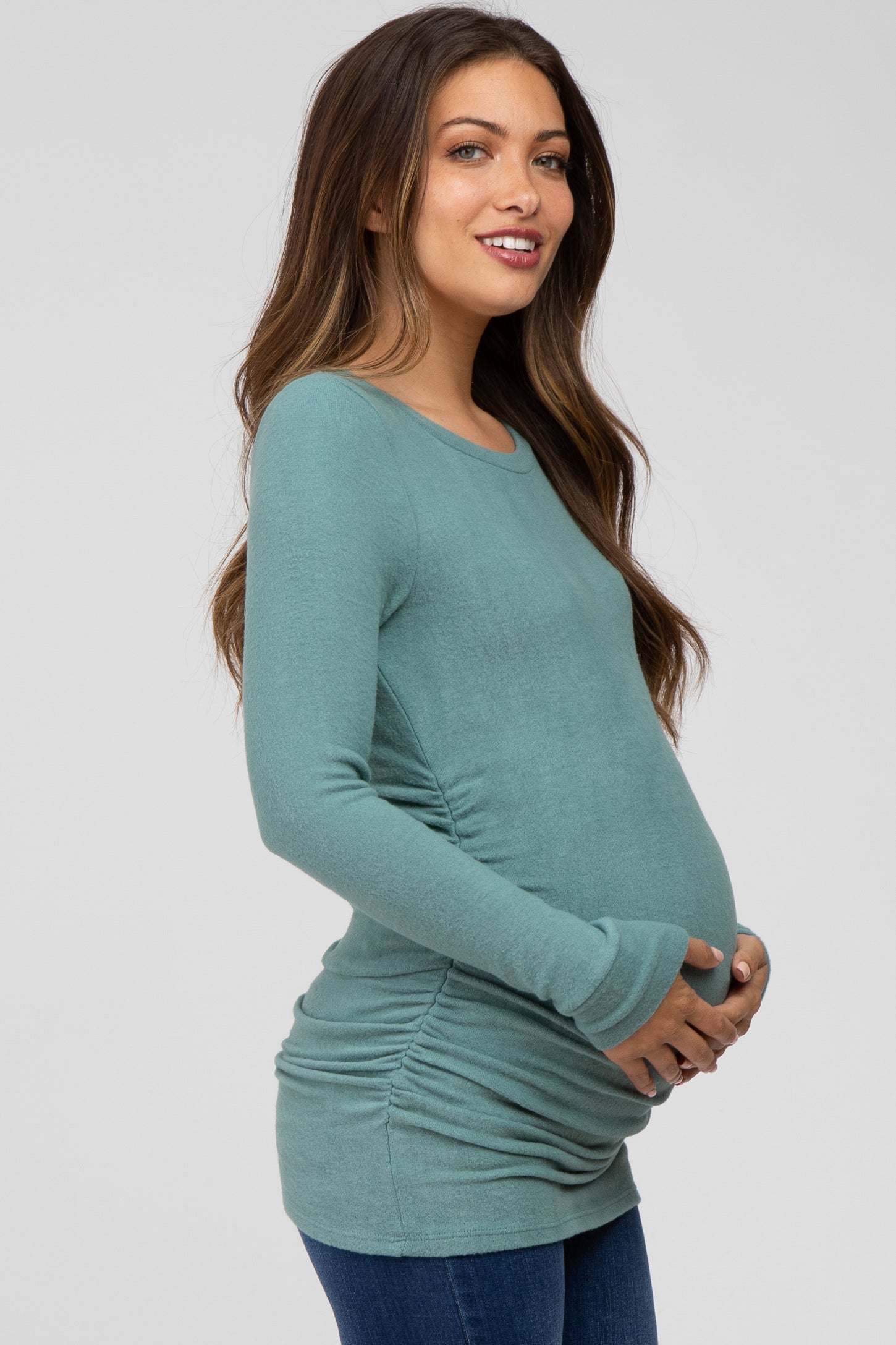 woman wearing a teal long sleeve maternity top with side ruching and holding her baby bump