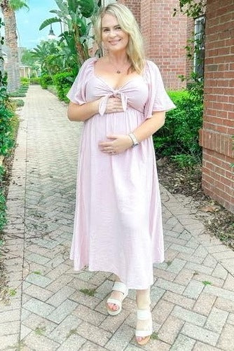 woman wearing a pink maternity dress with tie detail holding her baby bump outdoors