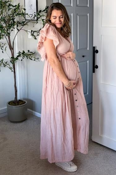 woman wearing a light pink maternity maxi dress with ruffled sleeves and buttons, holding her baby bump