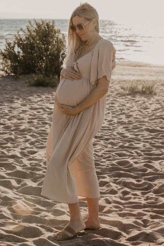 woman wearing a flowy beige maternity dress and holding her baby bump on a beach at sunset