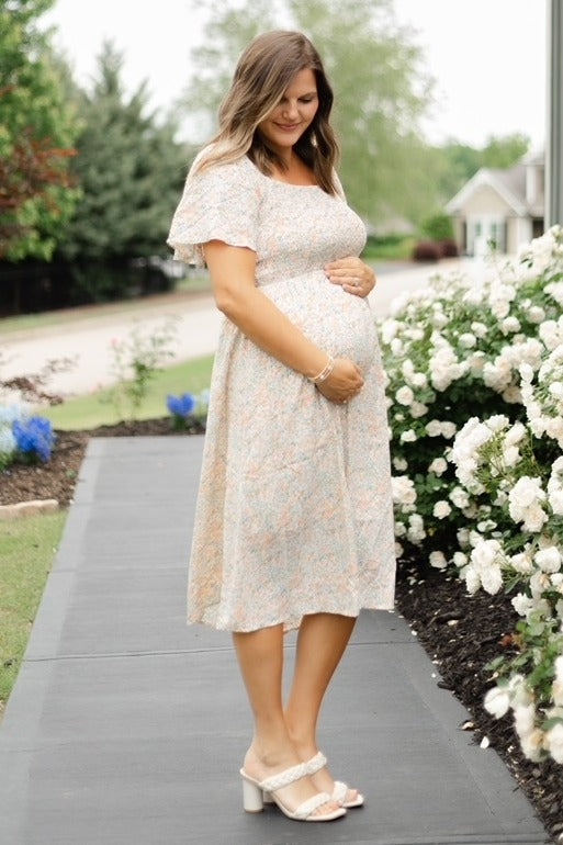 woman wearing a light floral maternity dress and holding her baby bump while standing outdoors