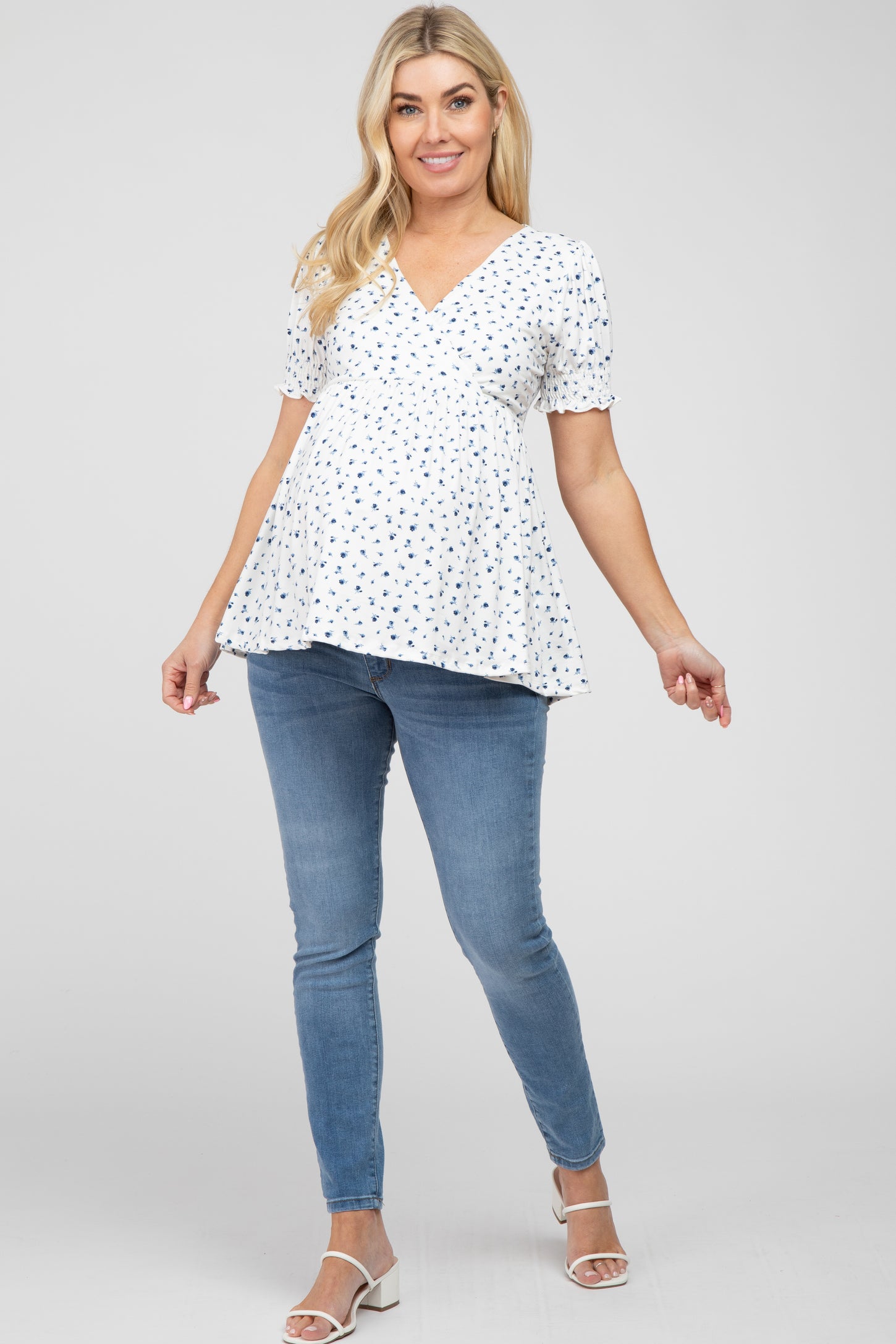 pregnant woman wearing a white floral maternity top and blue skinny jeans with white heels