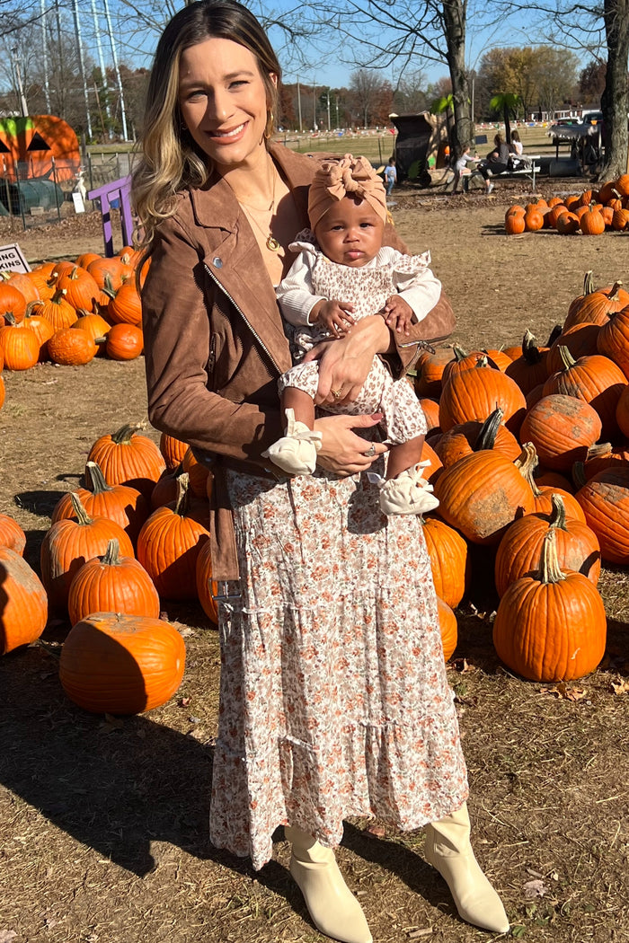 woman wearing a floral maxi dress and a brown jacket, holding a baby in a floral outfit among pumpkins