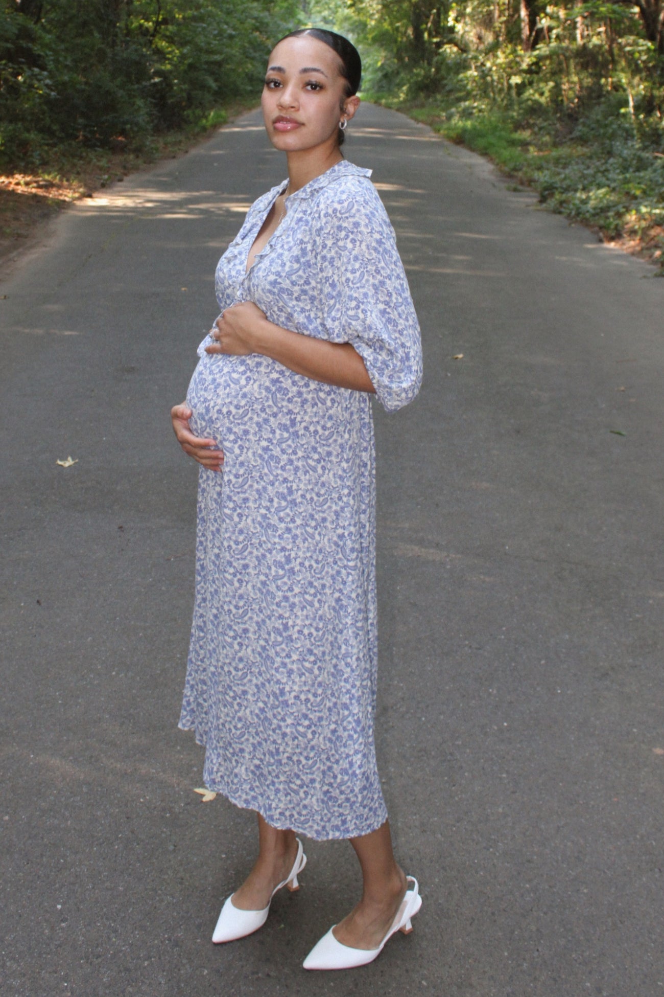 woman wearing a blue floral print maternity dress and holding her baby bump on a path in the woods