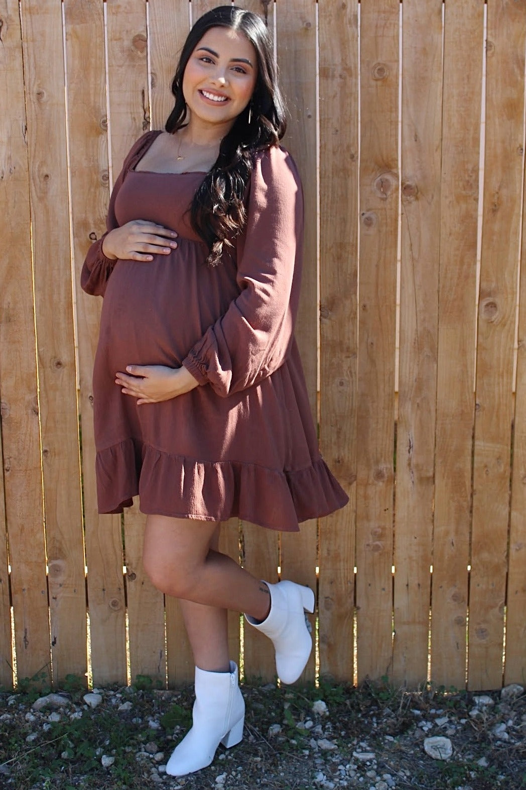Smiling pregnant woman in a mauve maternity dress and white boots posing against a wooden fence