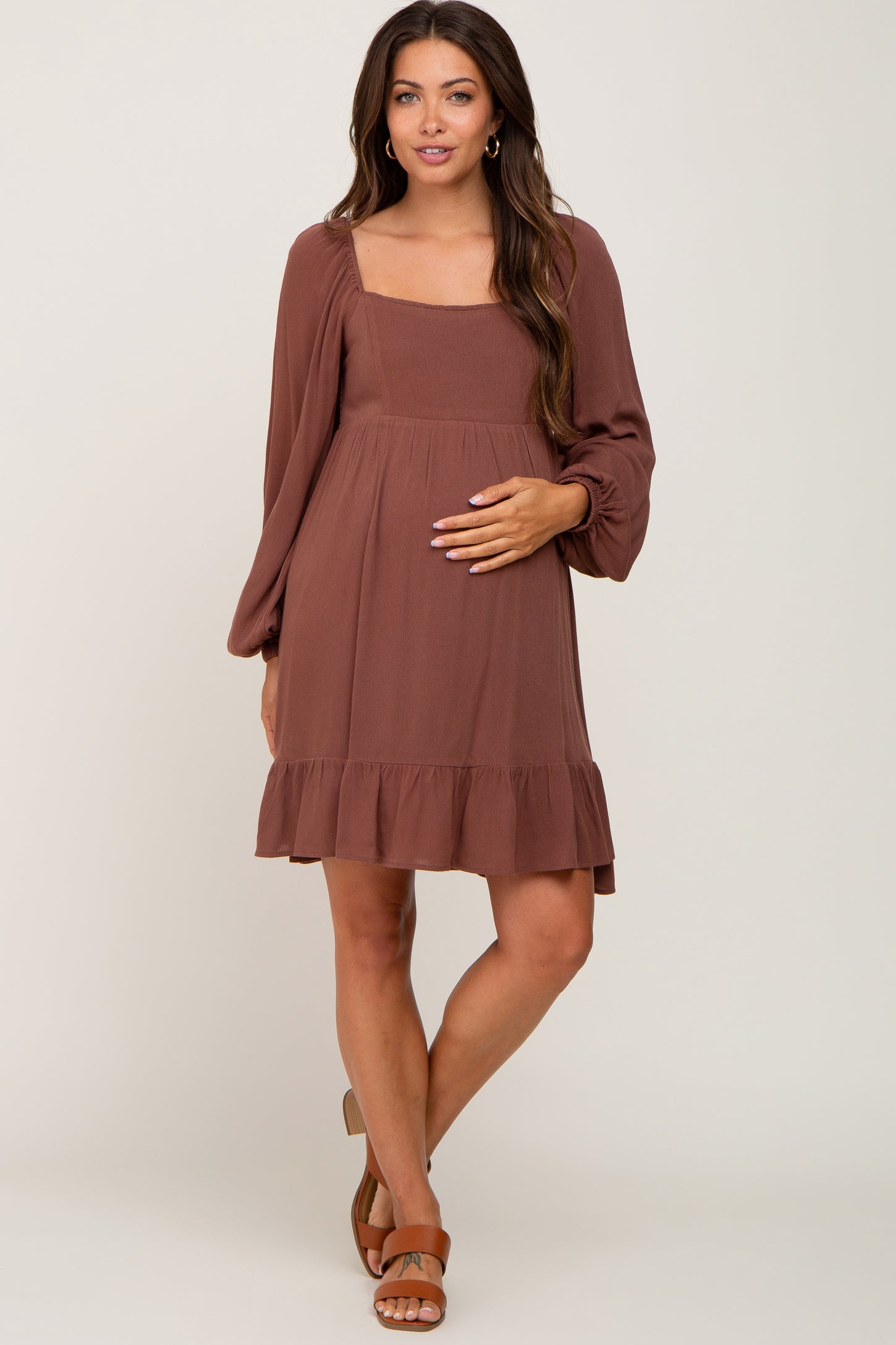 woman wearing a brown maternity dress with a square neckline, long sleeves, and ruffled hem, showing her baby bump