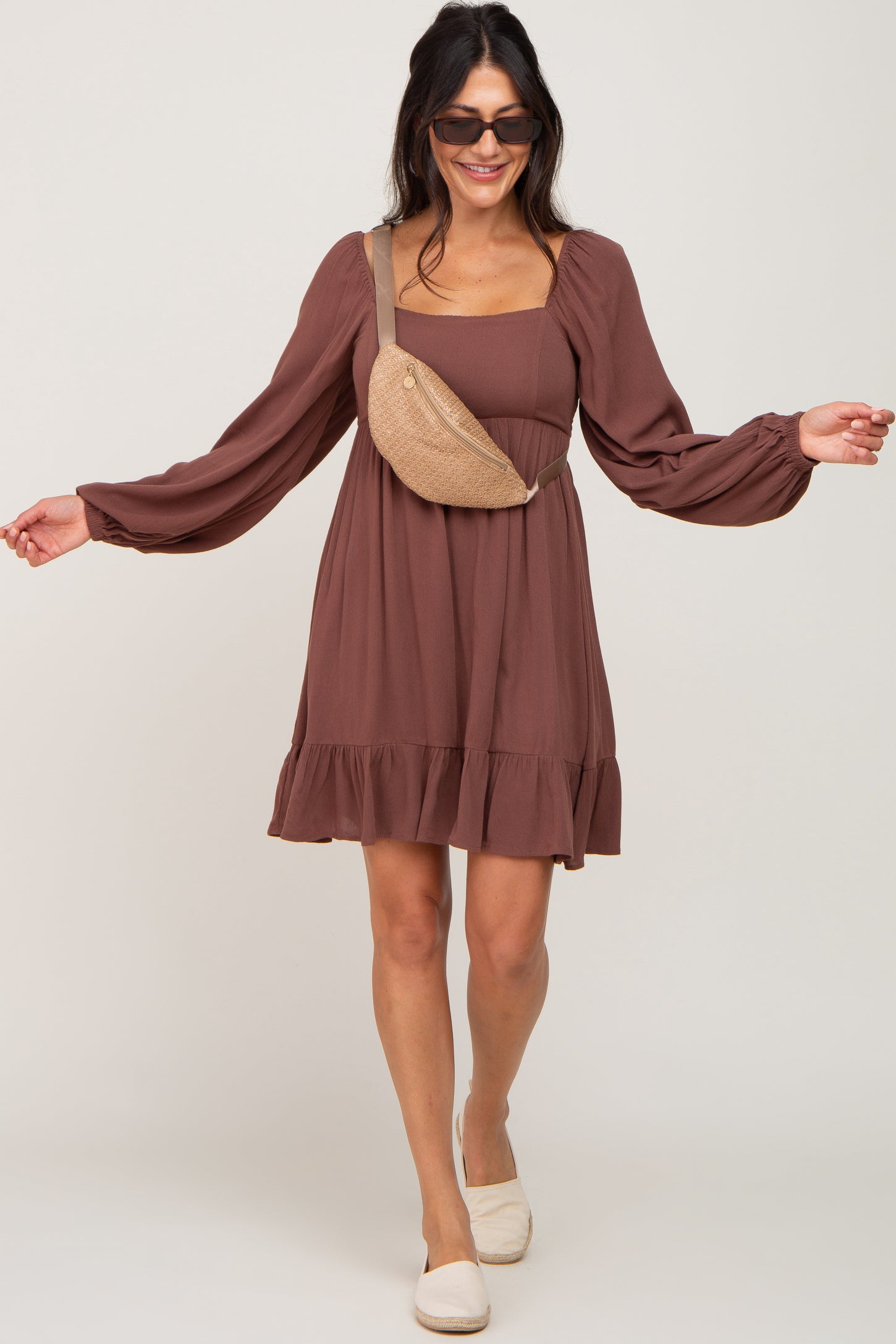 woman wearing a brown long sleeve mini dress with a ruffled hem and a tan fanny pack, styled with sunglasses