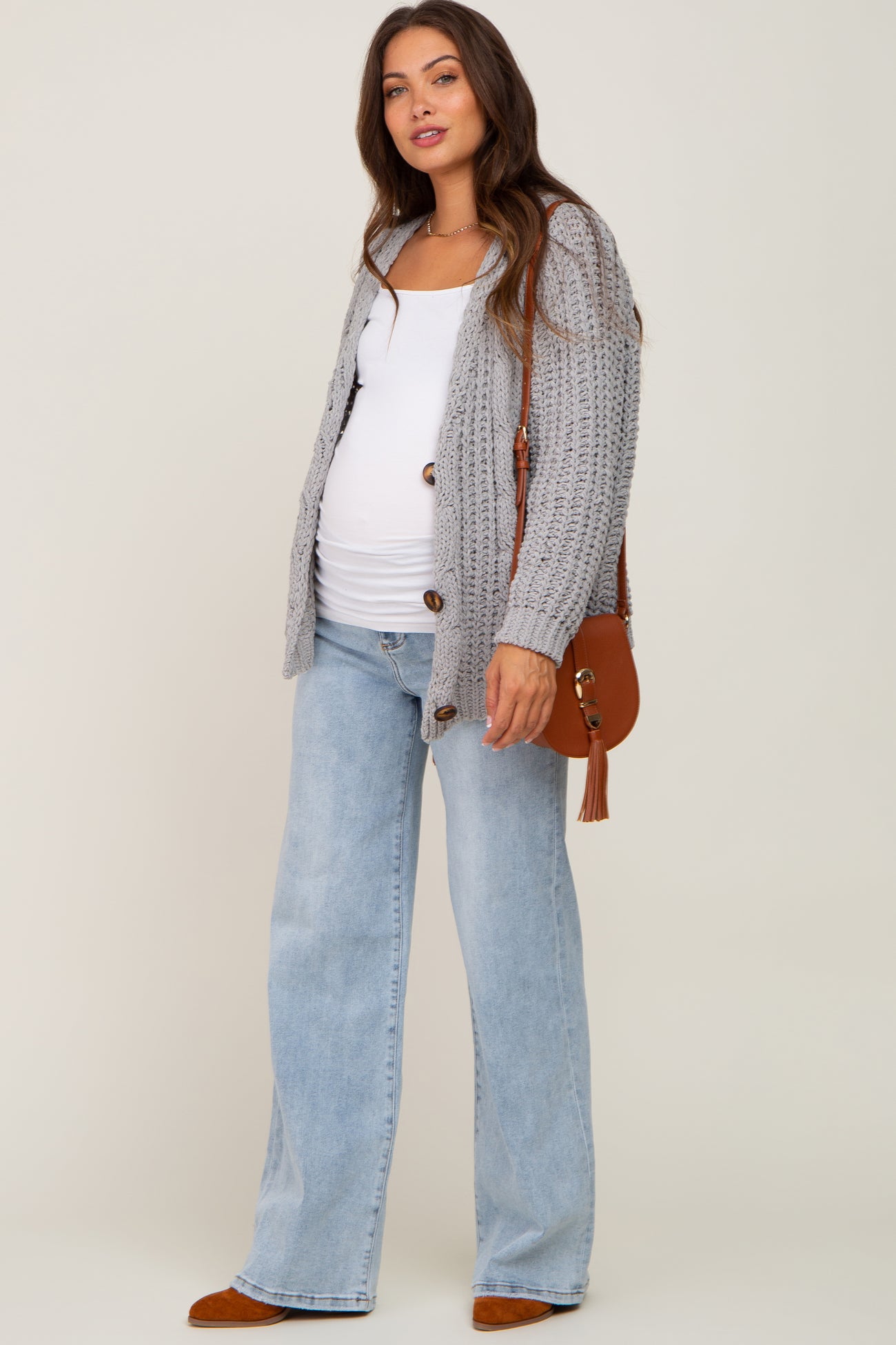 woman wearing a gray knit maternity cardigan over a white tank top and light wash wide-leg jeans, holding a brown handbag