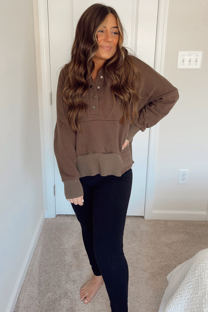 woman with long wavy brown hair wearing a brown quarter-button pullover sweatshirt and black leggings