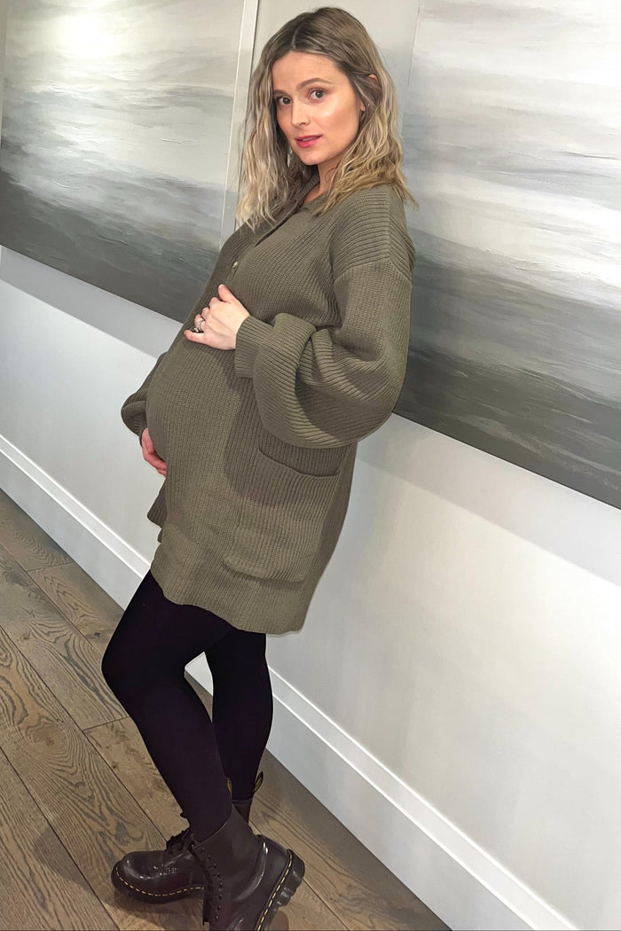 pregnant woman in olive green oversized cardigan and black leggings posing against a wall