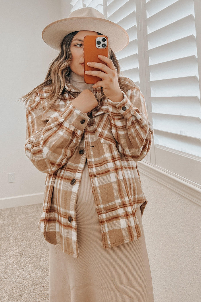 woman wearing a tan plaid shacket and wide brim hat taking a mirror selfie with an orange phone case