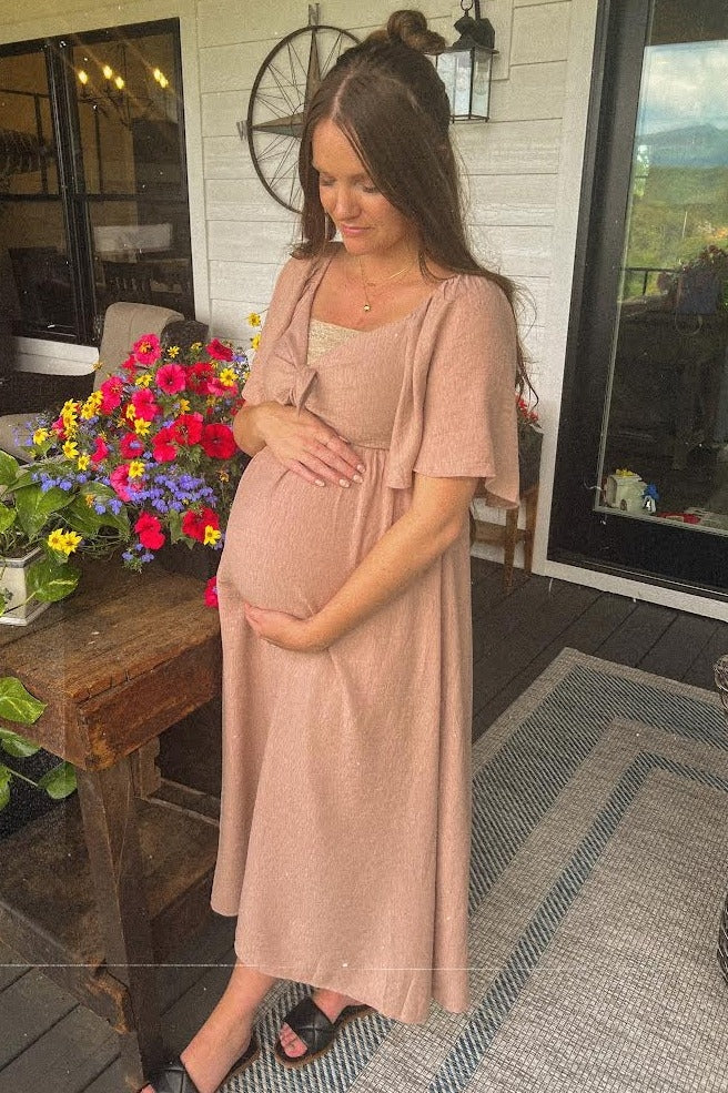 woman wearing a blush pink maternity dress with short sleeves and a cinched waist, cradling her baby bump outdoors