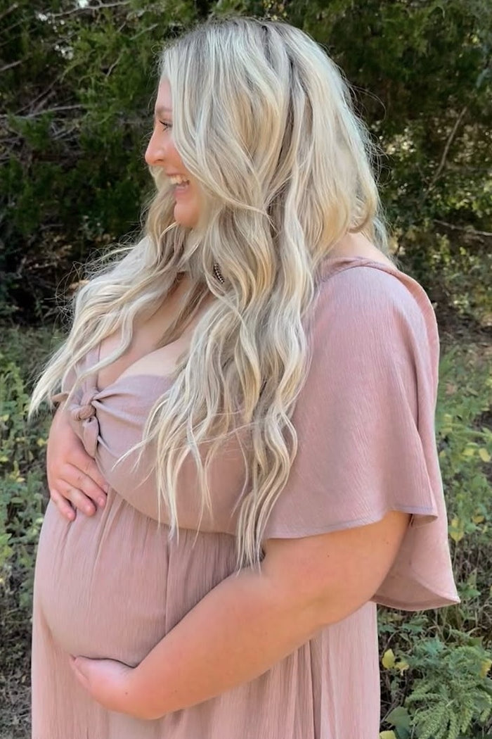 woman wearing a light pink maternity dress with a knot detail and holding her baby bump outdoors