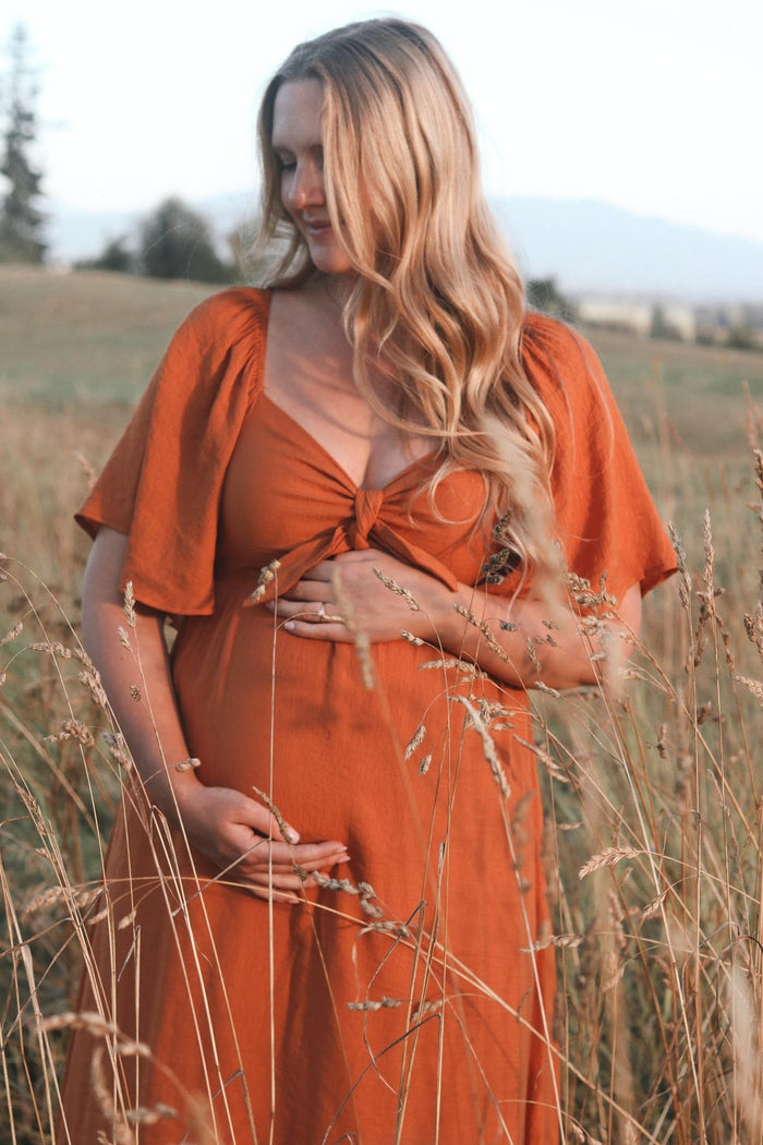 woman wearing an orange maternity dress with a knotted front, holding her baby bump in a field of tall grasses