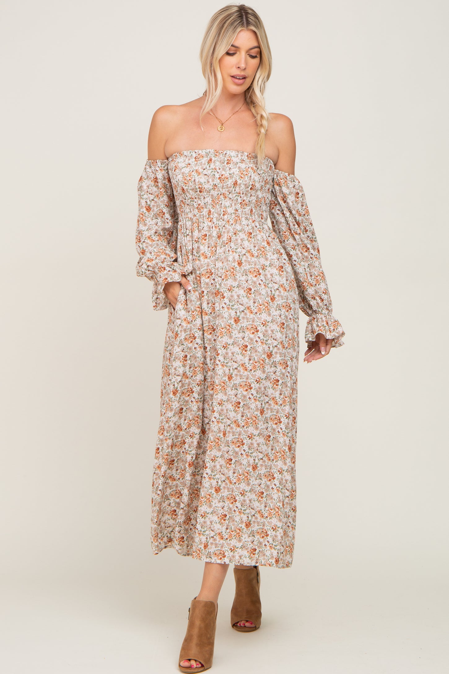 woman wearing a floral off-the-shoulder maxi dress with long sleeves and pockets, paired with tan heeled mules