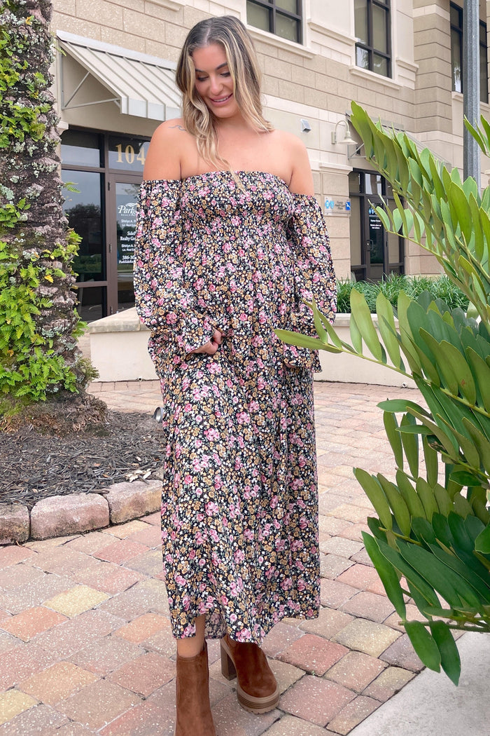 woman wearing a floral off-the-shoulder maternity maxi dress with a baby bump, standing outside with greenery around