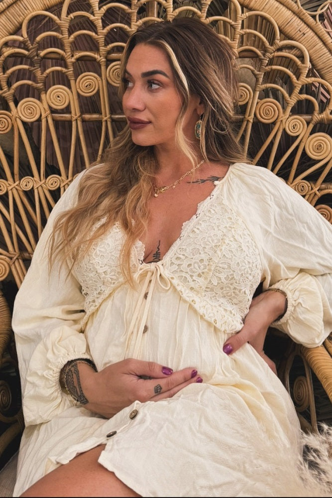 woman wearing a cream maternity dress with lace detail, sitting and cradling her baby bump in a rattan chair