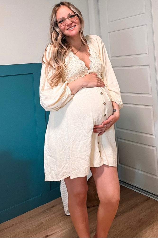 woman wearing a cream maternity dress with lace detailing and button front, holding her baby bump