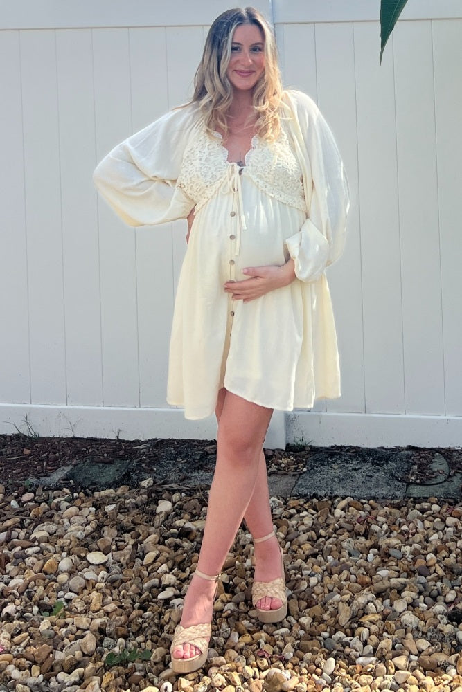 woman wearing a creamy white maternity dress with lace details and a baby bump, standing on a gravel surface