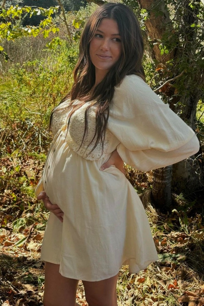 woman wearing a cream maternity dress with lace detailing, gently cradling her baby bump in a natural setting